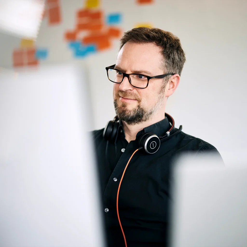 Mann mit Brille und Kopfhörern um den Hals arbeitet konzentriert an einem Computer. Im Hintergrund sind bunte Klebezettel an der Wand zu sehen.